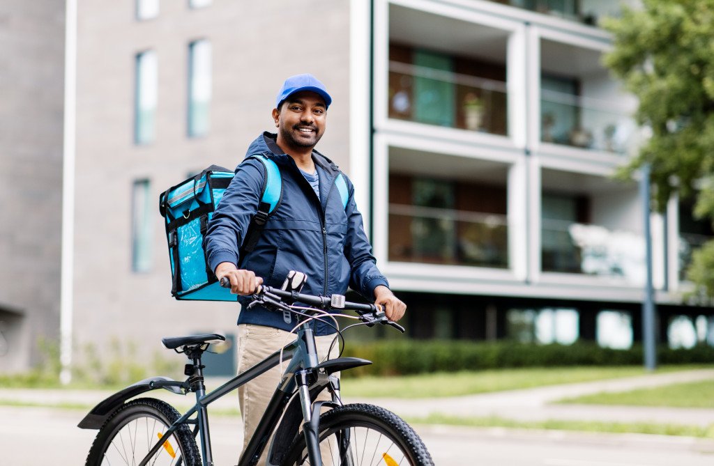 food shipping, profession and people concept - happy smiling indian delivery man with thermal insulated bag and bicycle on city street